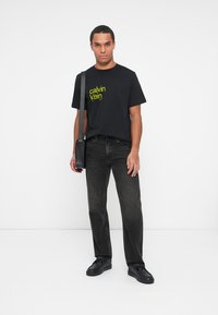 Juoda medvilnės marškinėliai su žaliu "Calvin Klein" logotipu, derinami su tamsiomis džinsinėmis kelnėmis ir juodais bateliais, dėvimi su juoda kryžminėmis maišeliu.