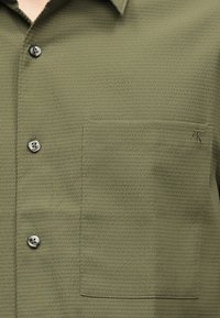 Πουκάμισο ελιάς με υφή, κουμπιά και αριστερή τσέπη στο στήθος με μικρή κεντημένη λογότυπο "cK".