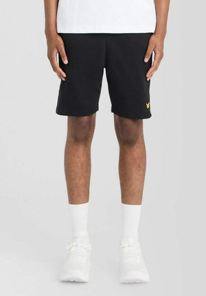 Mand iført sorte shorts med et lille gult logo, hvide sokker, hvide sneakers og en hvid skjorte mod en ensfarvet baggrund.