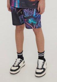 Pantaloni corti neri con grafiche astratte vivaci in blu, rosa e verde, caratterizzati da testi giocosi e dettagli di controller di gioco. Abbinati a sneakers bianche e nere.