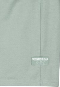 Mintgrünes T-Shirt aus strukturiertem Stoff, mit einem rechteckigen weißen Etikett am unteren Rand, das "DOWNTOWN CLUB" in fetter Schrift zeigt.