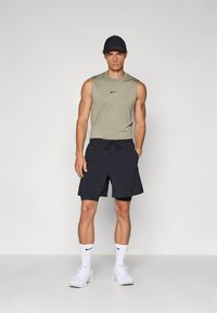 Hombre con gorra negra, camiseta sin mangas verde oliva, pantalones cortos negros, calcetines blancos de Nike y zapatillas blancas, de pie frente a un fondo sencillo.