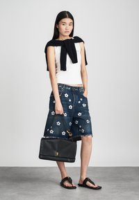 Denim shorts met bloemenborduursel, gedragen met een witte, aansluitende tanktop. Een marineblauwe gebreide trui hangt over de schouders. Zwarte handtas en sandalen.