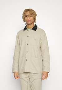 Beige Canvasjacke mit einem schwarzen Cordkragen, einer aufgesetzten Brusttasche und zwei unteren Taschen, mit Knopfverschluss und sichtbaren Nähdetails.