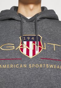 Felpa grigia con cappuccio e lacci, caratterizzata da un audace stemma ricamato in rosso, bianco e oro. Il testo recita "GANT AMERICAN SPORTSWEAR."