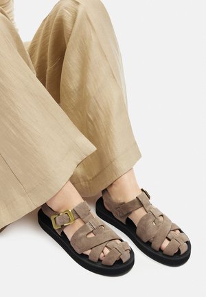 Braune Wildledersandalen mit überkreuzten Riemen, flacher schwarzer Sohle und einem metallischen Schnallenakzent an der Seite. Strukturierte Details auf den Riemen.