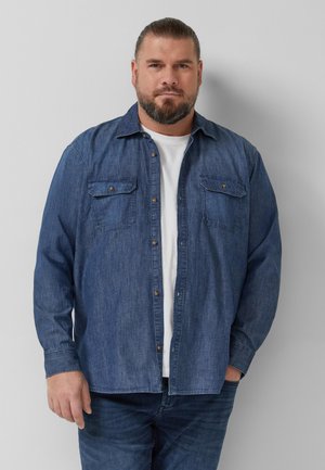 Homme avec barbe et cheveux attachés portant une chemise en jean déboutonnée sur un t-shirt blanc, debout devant un mur clair uni.