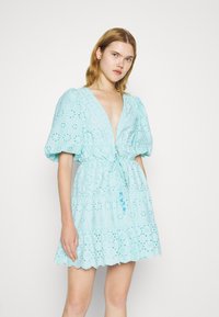 Robe bleu clair avec manches bouffantes, décolleté en V profond, broderie à œillets et jupe à plusieurs niveaux. Taille ajustable avec cordon de serrage et détail à pompon.