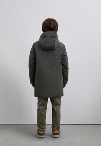 Donkergroene parka met capuchon, met een gladde buitenkant, rechte snit en zijnaden. Gedragen over lichtbruine broek en grijze sneakers.