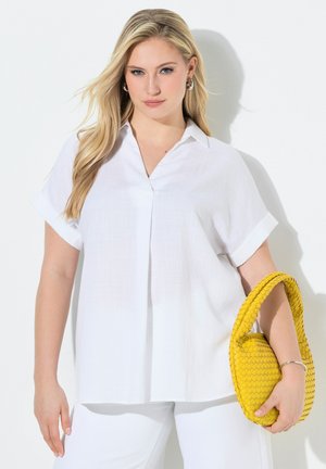 Blondynka w białej bluzce z krótkim rękawem i spodniach, trzymająca żółtą plecioną torebkę na białym tle.