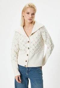 Strickcardigan in Off-White mit einem breiten Kragen, Knopfverschluss und strukturierten Mustern. Mit gerippten Bündchen und Saum, sowie goldfarbenen Knöpfen.