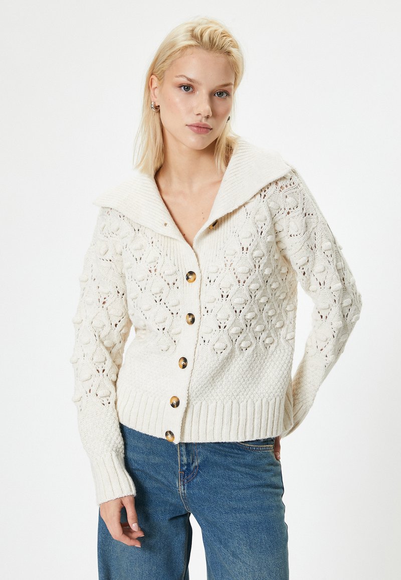 Gebreide cardigan in off-white met een brede kraag, knoopsluiting en textuurpatronen. Voorzien van geribbelde boorden en zoom, met goudkleurige knopen.