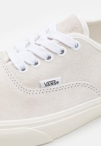 Baskets basses en daim beige avec un design à lacets blancs, œillets ronds et petit logo Vans noir près de la couture latérale. Semelle en caoutchouc.