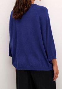 Frau mit braunen Haaren, die einen locker sitzenden, langärmligen blauen Pullover und schwarze Hosen trägt, von hinten vor einem hellen Hintergrund dargestellt.