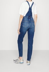 Jeanslatzhosen mit verstellbaren Schultern, schmalen Beinen und zwei Gesäßtaschen. Über einem weißen T-Shirt getragen, kombiniert mit silbernen Sneakers.