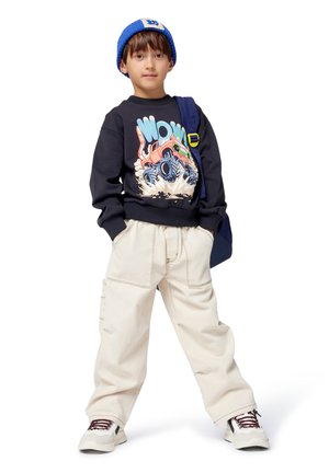 Ung gutt som har på seg en blå lue, svart hettegenser med monstertruckgrafikk, beige vide bukser, hvite sneakers og bærer en marineblå ryggsekk.