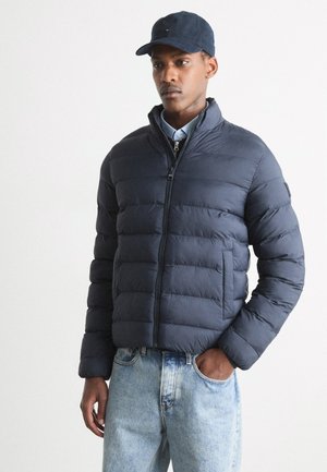 Giacca puffer blu navy con zip frontale, colletto alto e texture trapuntata, abbinata a jeans di denim blu chiaro. Cappellino nero indossato in testa.