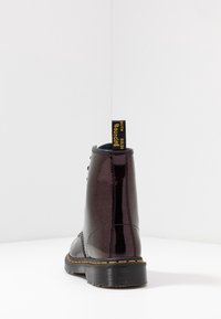 Botín de cuero sintético negro y púrpura con cordones negros, tirador con logo amarillo y suela de goma negra con costuras visibles.