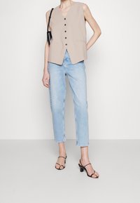 Chaleco sin mangas color beige con escote en V y seis botones oscuros, combinado con pantalones vaqueros rectos de color azul claro y sandalias de tacón negras con tiras.