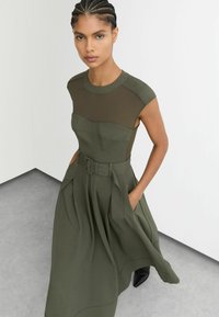 Oliivrohelist värvi kleit, millel on lühikesed nokkamisega varrukad, vööga istuv ülakeha ja hõlmikseelik taskutega ning pehme kangatekstuur.