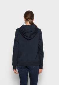 Donna con i capelli castani raccolti in una coda di cavallo, che indossa un hoodie nero e jeans scuri, in piedi di fronte a uno sfondo grigio chiaro uniforme.