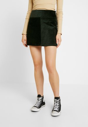 Kobieta ubrana w mini spódniczkę z zielonego sztruksu, beżową bluzkę z długim rękawem i czarne trampki za kostkę z białymi sznurowadłami na białym tle.