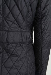 Casaco acolchoado preto com um padrão em diamante, textura suave, detalhe de botão e um corte ajustado com uma cintura ligeiramente afunilada.