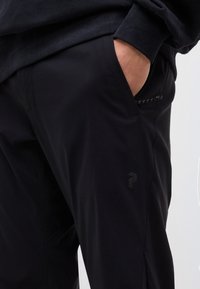 Personne portant un pantalon noir et un haut à manches longues foncé, la main droite reposant dans la poche avant, sur un fond clair uni.