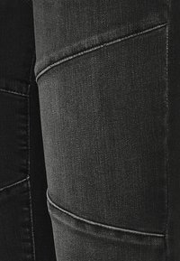 Gros plan de jeans en denim noir montrant les coutures détaillées et la texture sur les zones de la cuisse et du genou.