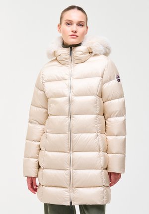 Vrouw draagt een lichtbeige donsjas met een met bont afgezoomde kap, staand tegen een effen lichte achtergrond.