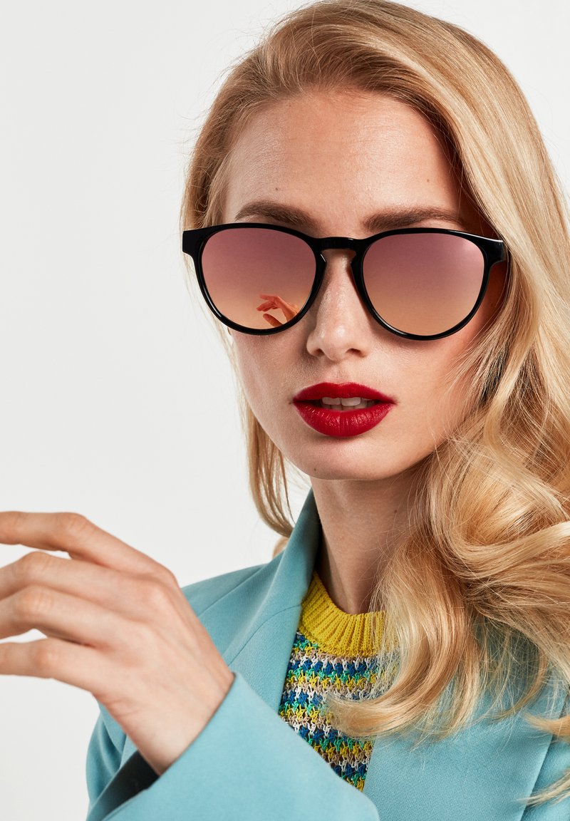 Ochelari de soare tip cat-eye negri cu lentile gradiente roz. Purtați de o persoană cu păr blond, îmbrăcată într-o jachetă bleu deschis peste un pulover multicolor.