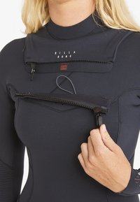 Person pulling open chest zipper of black Billabong wetsuit showing neckline and stitching details.