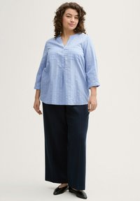 Hellblaue Bluse mit langen Ärmeln, V-Ausschnitt und zwei Brusttaschen, kombiniert mit weit geschnittenen, navy-blau gestreiften Hosen und schwarzen Slipschuhen.