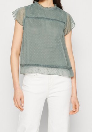 Vrouw draagt een doorschijnende, kortärmelige blouse in saliegroen met een gestippelde textuur en een gerimpelde kraag, gecombineerd met witte, high-waisted broek.