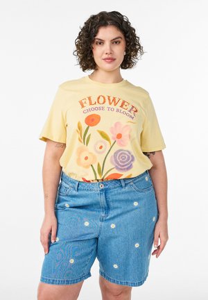 Kvinde med krøllet hår iført en gul t-shirt med blomsterprint og denimshorts med små broderede margueritter, stående foran en hvid baggrund.