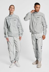 Lysegrå træningsdragter lavet af letvægtsstof, med en lynlåstrøje og elastiske manchetter, med kontrasterende mørke detaljer og logo.