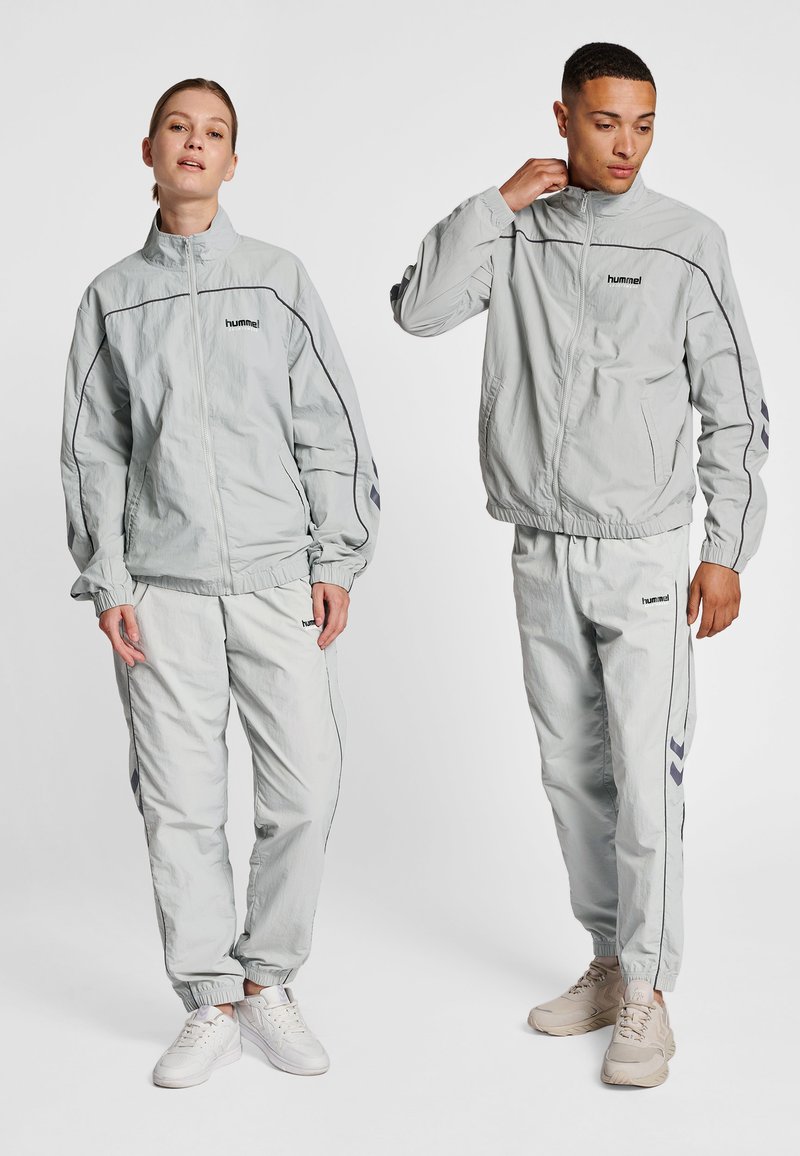 Lysegrå træningsdragter lavet af letvægtsstof, med en lynlåstrøje og elastiske manchetter, med kontrasterende mørke detaljer og logo.