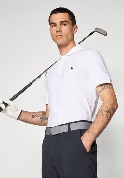 Valkoinen polo-paita, jossa on lyhyet hihat ja laivastonsininen logo, yhdistetty tummiin housuihin ja harmaaseen vyöhön. Golf-maila pidetään olalla ja kädessä on hansikas.