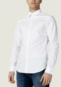 Camicia bianca a maniche lunghe in cotone con colletto a punta, dotata di un logo discreto e orlo dritto; abbinata a jeans blu.