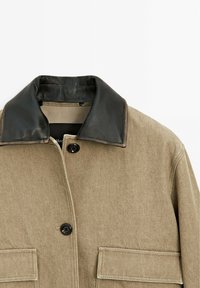 Manteau beige avec col en cuir noir, deux poches avant à rabat et boutons noirs sur un fond blanc uni.