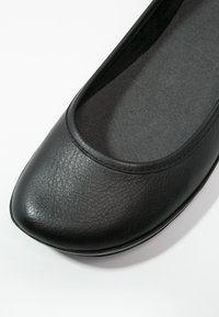 Chaussure plate en cuir noir avec un bout carré arrondi et une texture lisse, montrant le dessus et la doublure intérieure sur fond blanc.