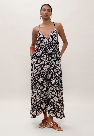 Mujer de pie con un vestido maxi negro con flores y tirantes finos, pulsera de cuentas rojas, pendientes de aro dorados y sandalias con flores rojas.