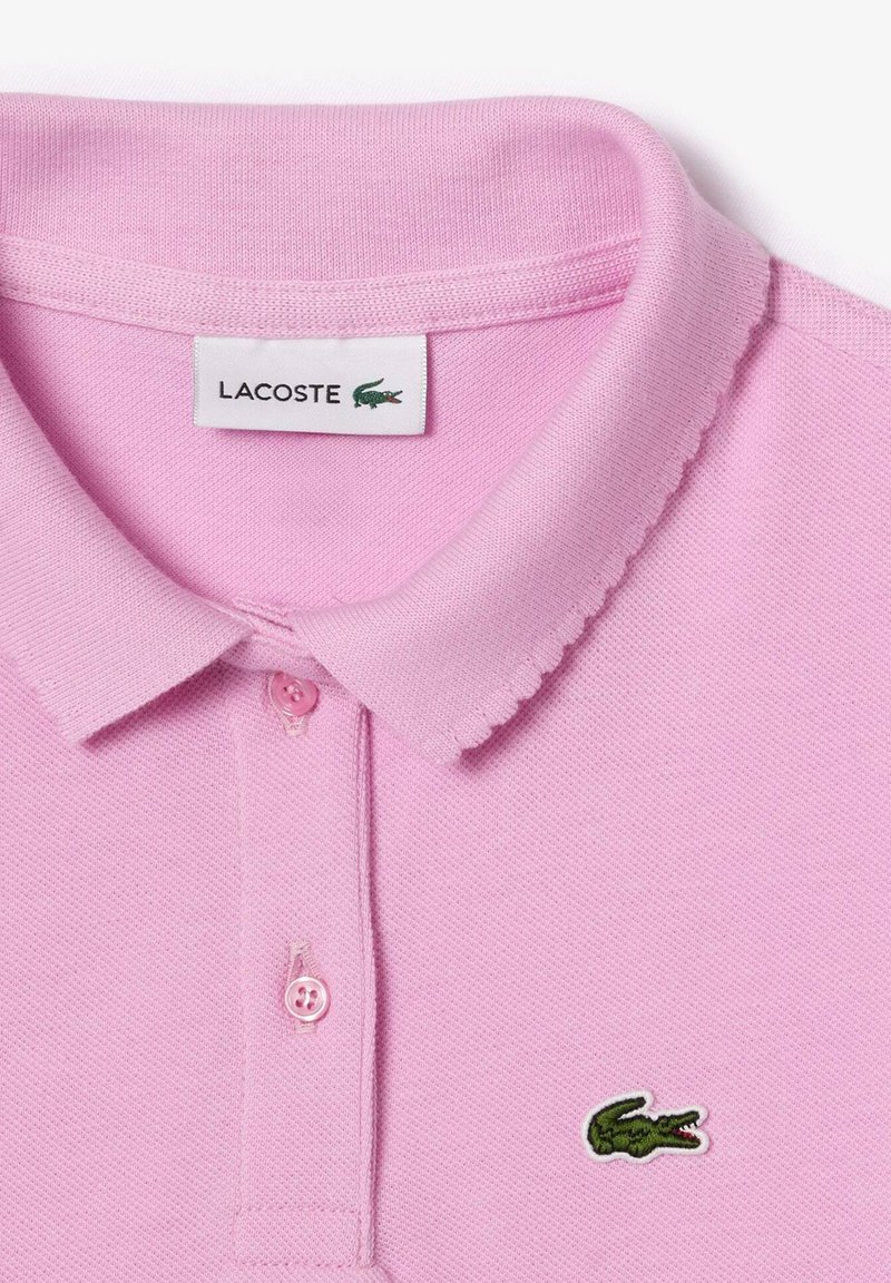 Polo rose clair en tissu texturé, avec un col, une patte de boutonnage à trois boutons et un logo de crocodile vert brodé.