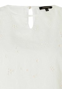 Weiße Bluse mit floralem Stickmuster. Detail mit Schlitz am Rücken, kurze Ärmel, leichtes Material, strukturierte Oberfläche mit erhabenen Blumenmotiven.