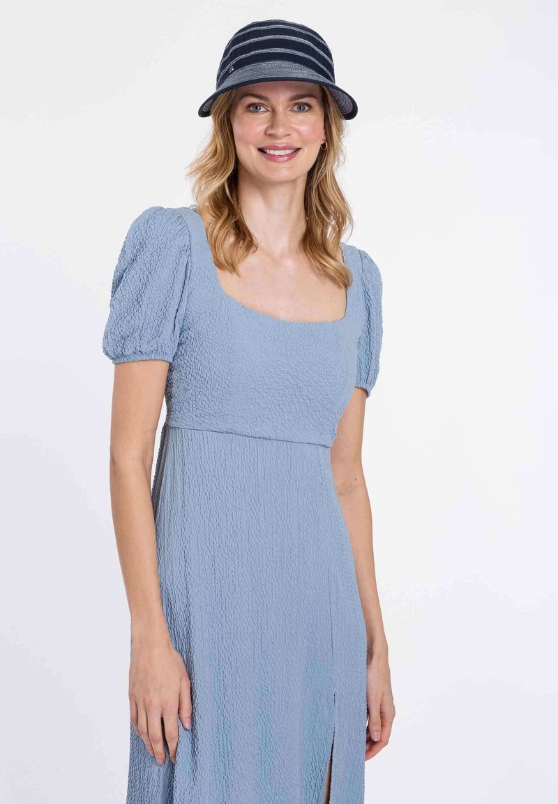 Šviesiai mėlyna suknelė su tekstūrizuota medžiaga, pūstomis rankovėmis ir kvadratiniu iškirpimu, su šoniniu plyšiu. Derinama su tamsiai mėlynu raštuotu skrybėle.