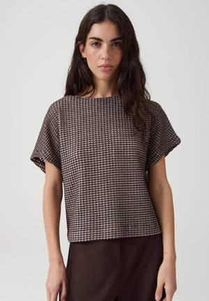 Donna con lunghi capelli scuri indossa una maglietta marrone a maniche corte con motivo e pantaloni scuri su uno sfondo chiaro uniforme.