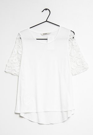 Blusa blanca con cuello redondo y mangas cortas de encaje colgada en una percha negra contra un fondo blanco.