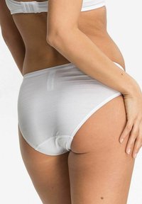 Frau in weißer Unterwäsche und weißem BH, von hinten und von der Seite gezeigt, mit einer Hand auf der Hüfte vor einem schlichten Hintergrund.