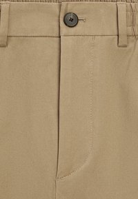 Béžové cargo kalhoty s knoflíkovým zapínáním, přední kapsou a rovnými nohavicemi. Látka má měkký, strukturovaný povrch s viditelným stehováním.