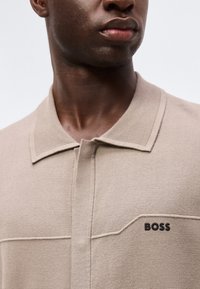 Κοντινό πλάνο άνδρα που φοράει μπεζ πλεκτό πόλο με ριμπ κολάρο και το λογότυπο «BOSS» στο αριστερό στήθος.